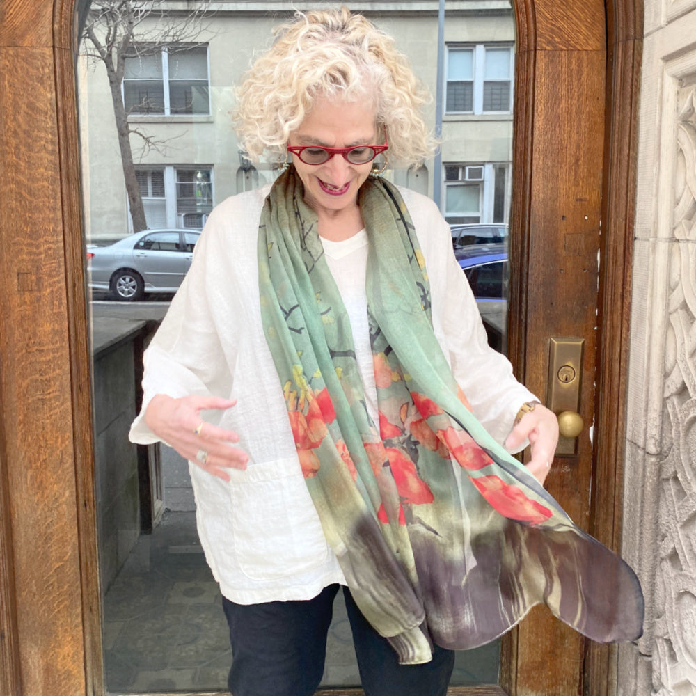 Senior woman wearing a pretty silk scarf over top a loose fit white linen top. She is standing outside and it is a windy day.
