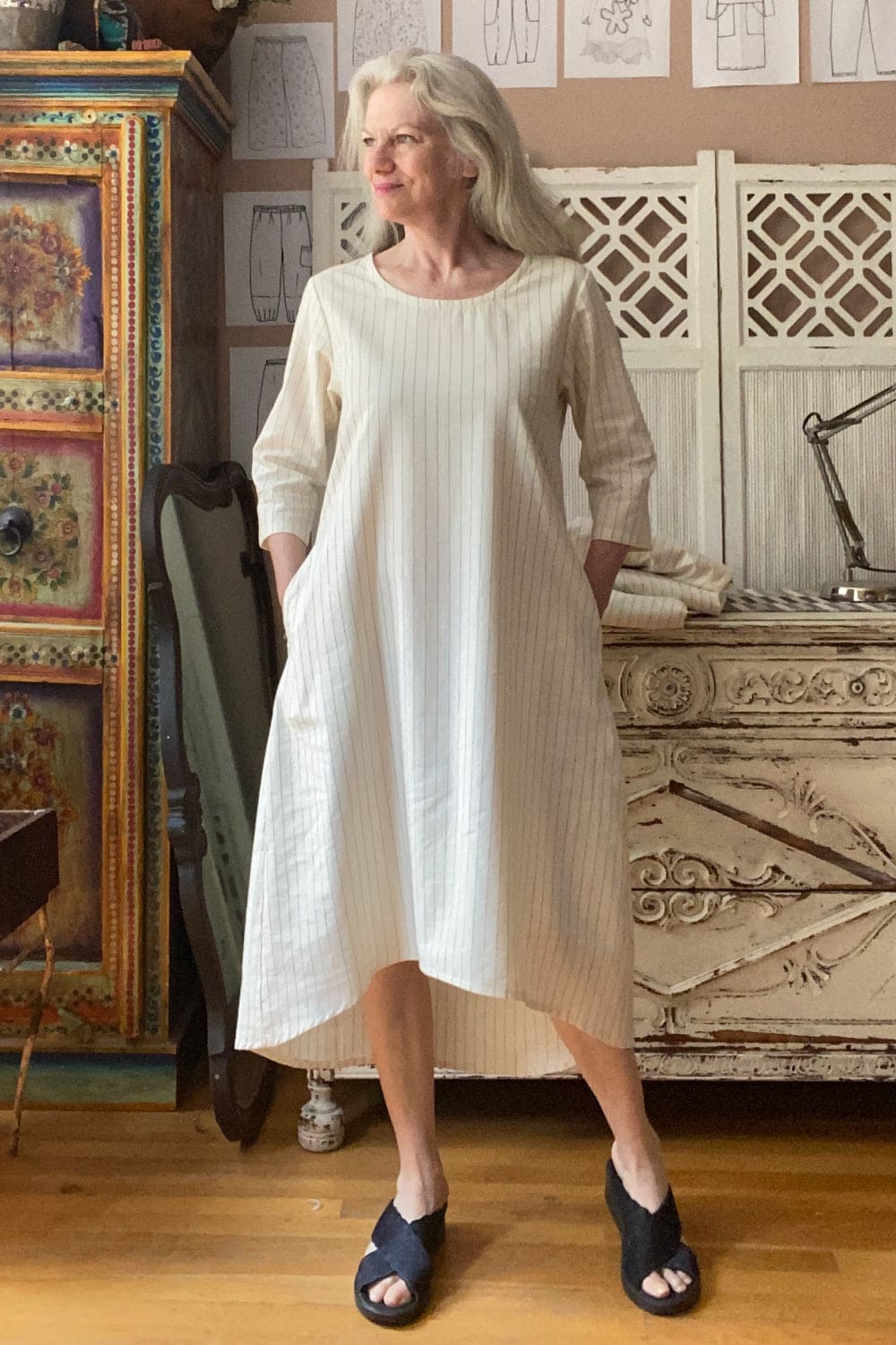 Creme color linen dress with a round neckline and curved hem.  Older woman with grey hair wearing also black sandles.