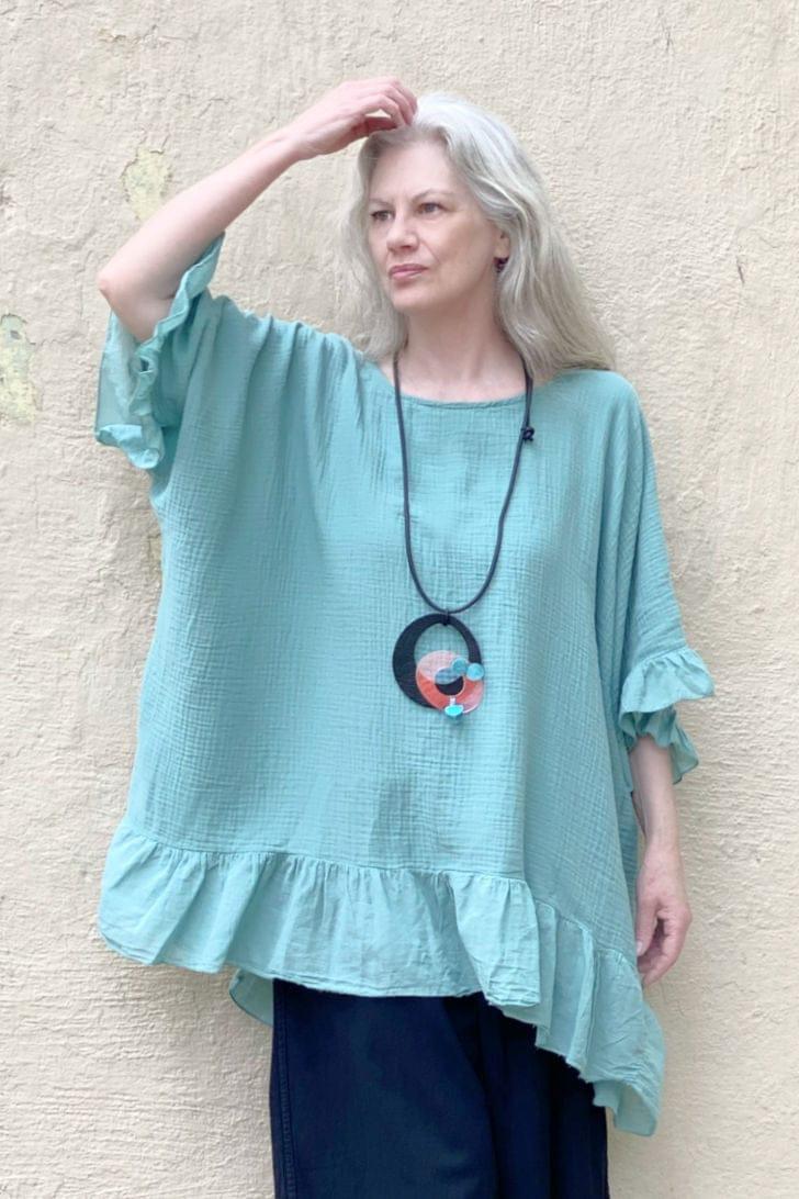 Woman wearing a pendant designed with circles hung on a black cord. She is wearing a ruffled cotton turquiose tunic with a black full cut pair of pants