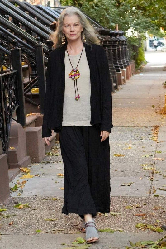 Older woman with long grey hair walking on a street wearing a black open cardigan jacket and black full cut black pants. She is also wearing a light biege tee, colorful fabric pendant and beige seed earrings with matching sandles.