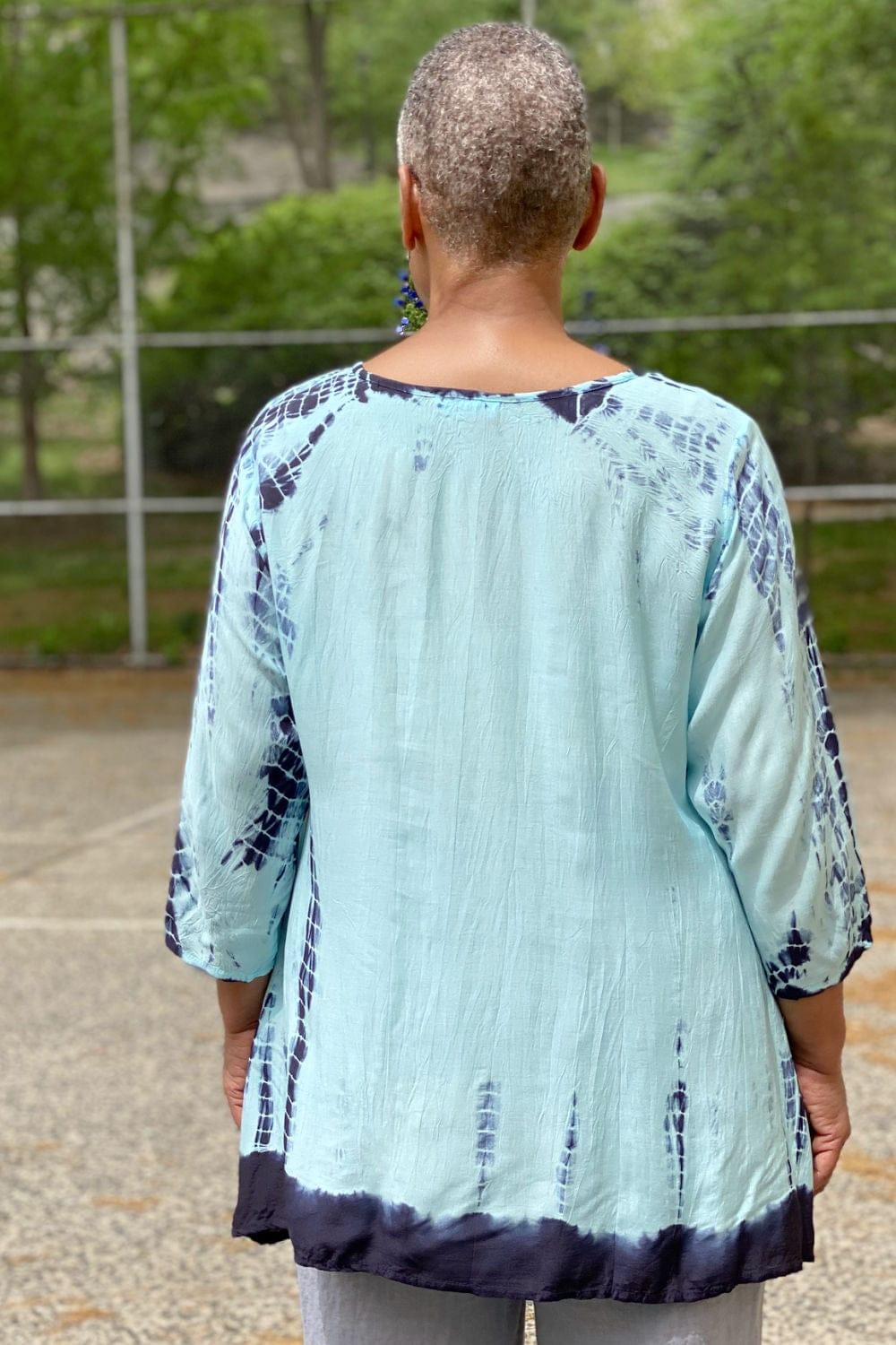 Back view of a Woman wearing a tie dye Aline shaped top in blue hues. She is also wearing dangle wooden earrings and natural color linen pants.