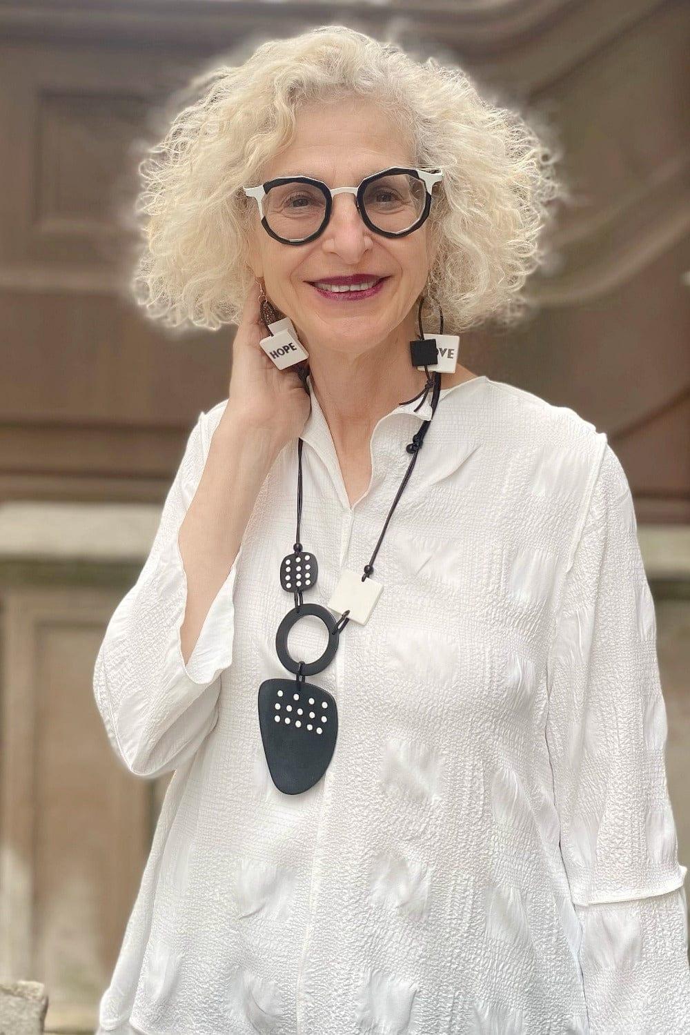 Smiling curly blond haired woman with funky glasses wearing a white blouse , black and white statement neckace and earring printed with the words hope and love,