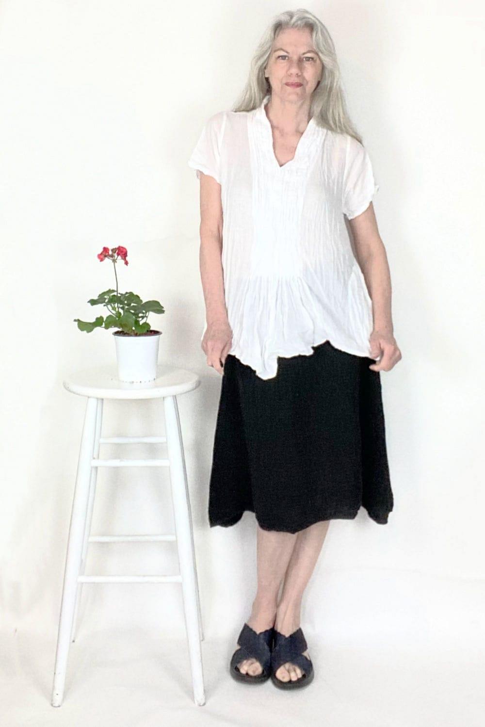 Woman with long grey hair wearing a short sleeve white cotton top with a aline cut cotton black skirt. 