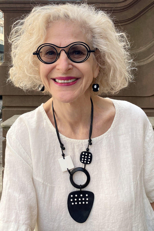 Smileing older woman with cropped blond hair wearing black rim glasses, linen white short sleeve top and Black with White Polka Dots Earrings shaped round and smooth surface with a French Hook. Also wearing a black and white graphic adjustable necklace with polka dots.