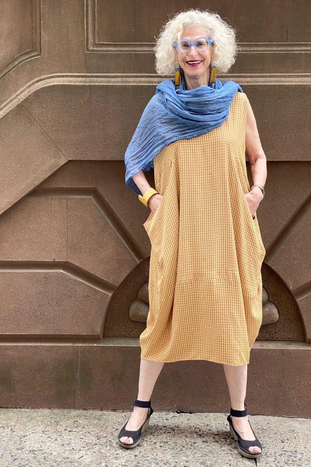Happy woman standing on the street wearing a summer dress with  a slight bubble shapem tank with two large front pockets. The fabric is a baby check pattern. She is also wearing a blue cotton scarf, blue and yellow tassel earrings with a yellow wooden bracelet.