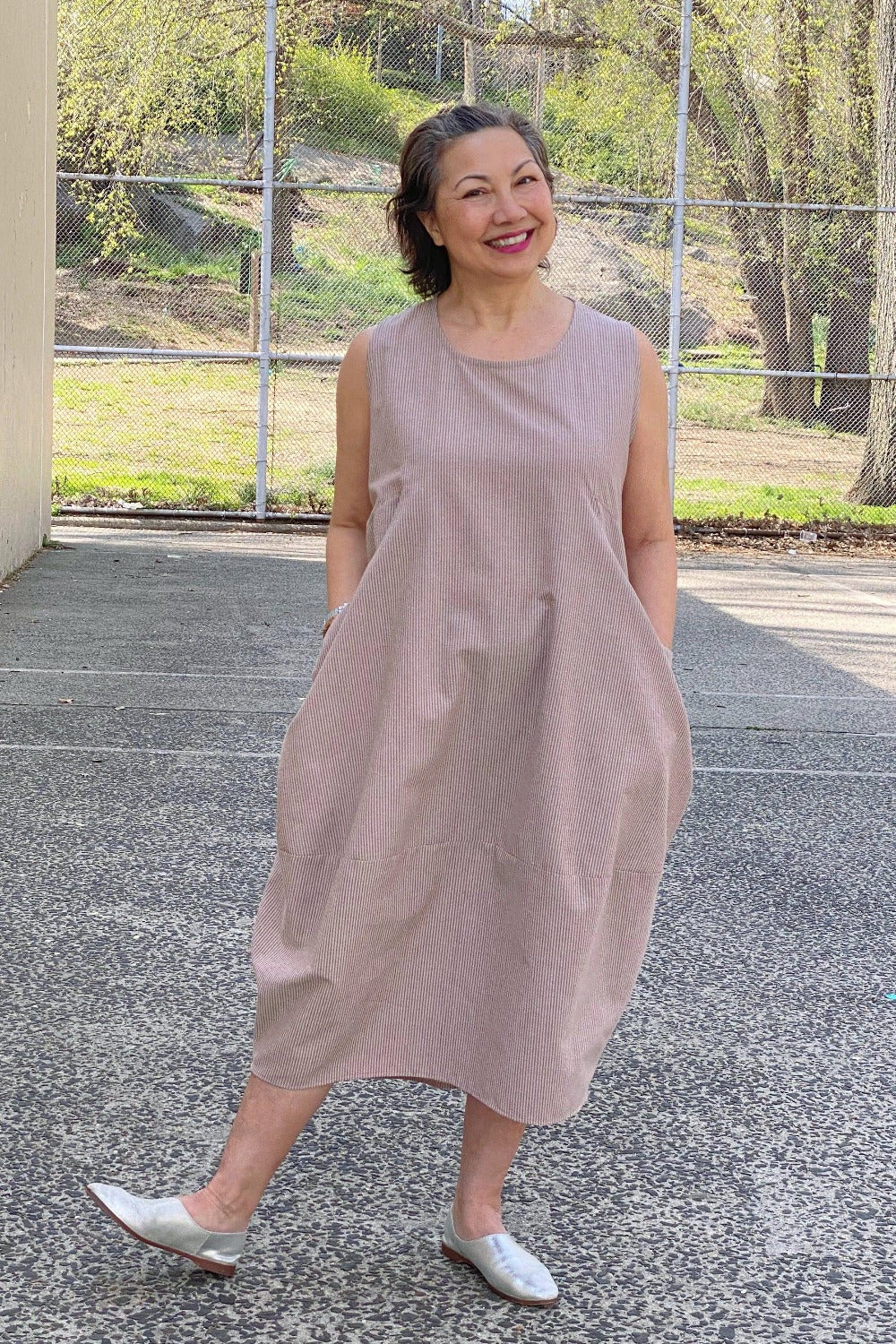 Smiling woman posing outsind wearing a tank dress with a slight bubble shape. It has 2 large front pockets and is a mocha colored seersucker fabric.