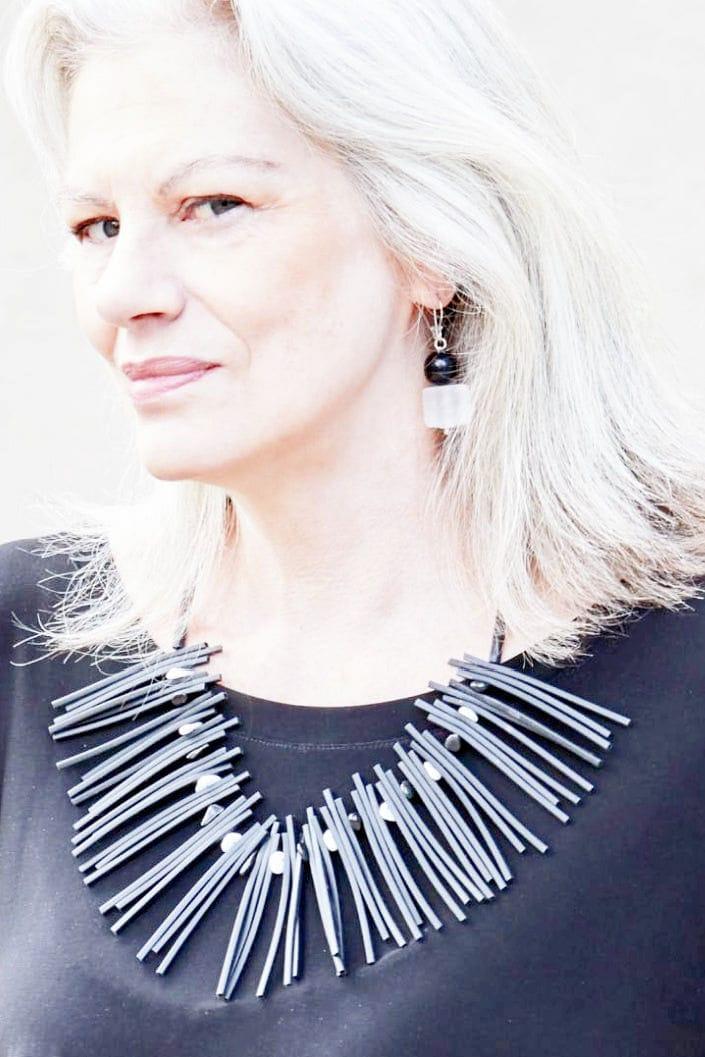 Black rubber necklace with beads worn by a grey hair woman. Black and clear beaded earings are worn as well. Necklace is worn over a black round neck top.