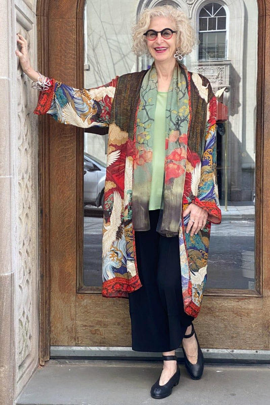Smiling woman over 60 years standing in a doorway. She is wearing a beautfful multicolored long open silk jacket. The print is asian inspired. She looks very stylish and is also wearing black full cut pants, a soft green top and a floral silk scarf.