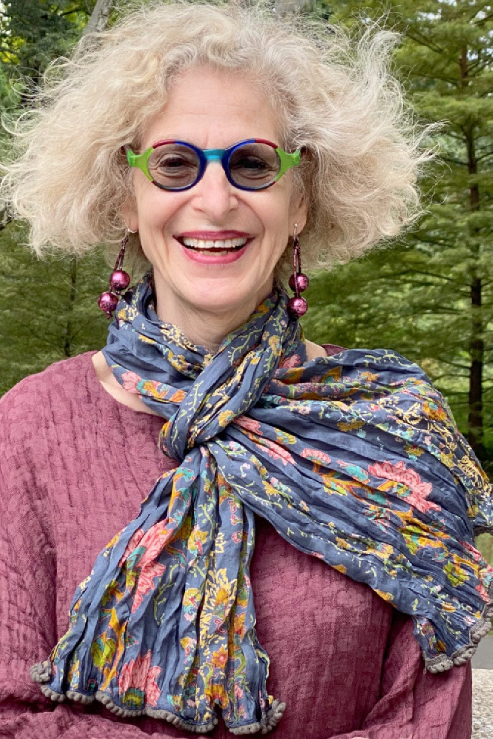 Women's funky earring made of two pink gilden balls hanging from leatherette cord. Smiling older woman wearing a rose colored textured jacket and floral cotton scarf.