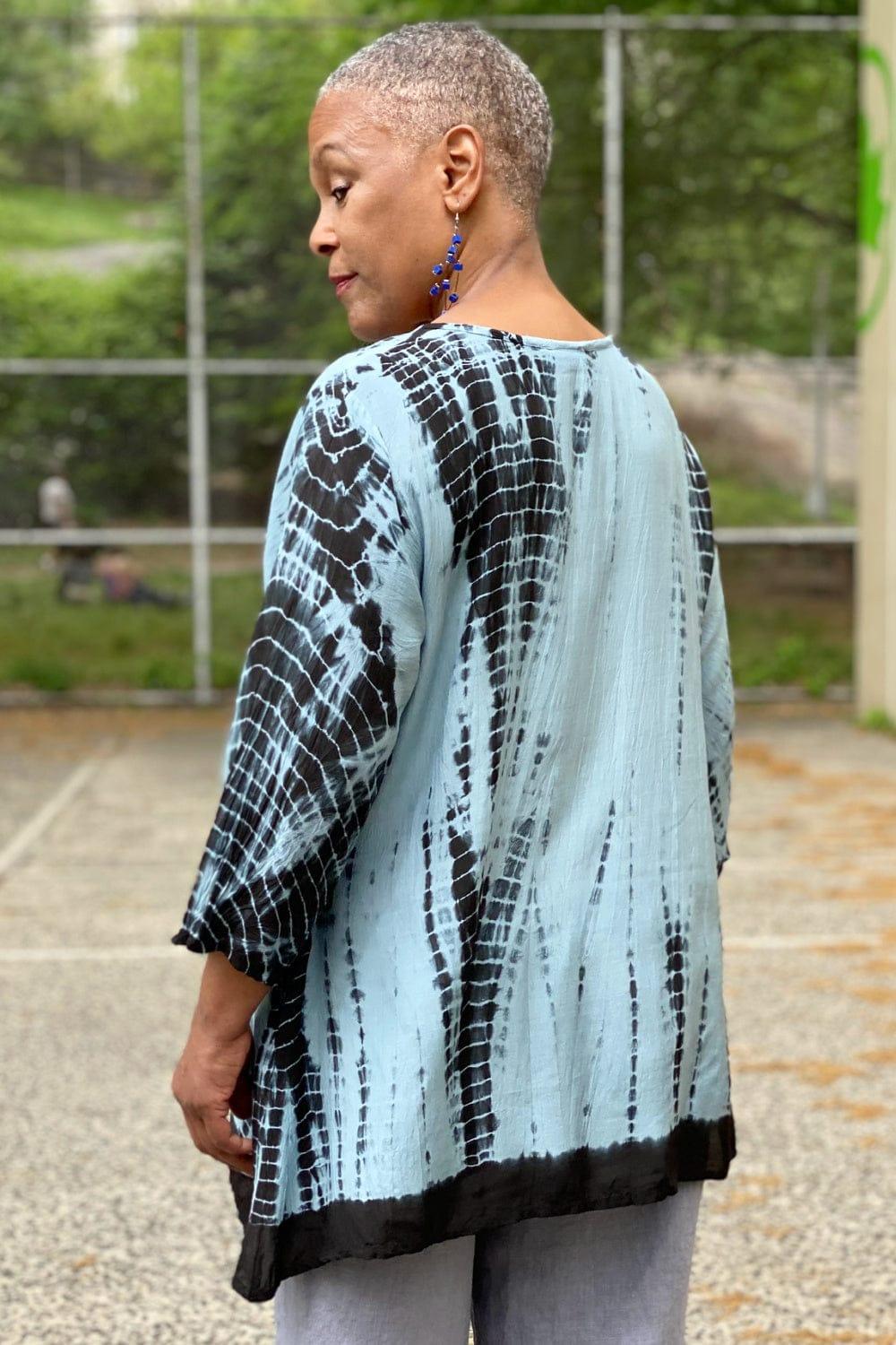 Back view of a Woman wearing a tie dye Aline shaped top in blue hues. She is also wearing dangle wooden earrings and natural color linen pants.