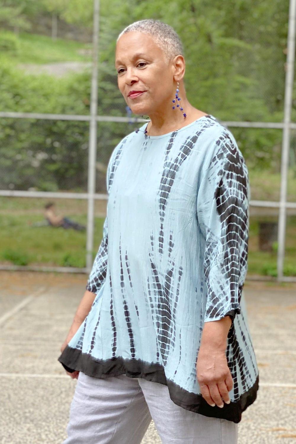 Woman wearing a tie dye Aline shaped top in blue hues. She is also wearing dangle wooden earrings and natural color linen pants.