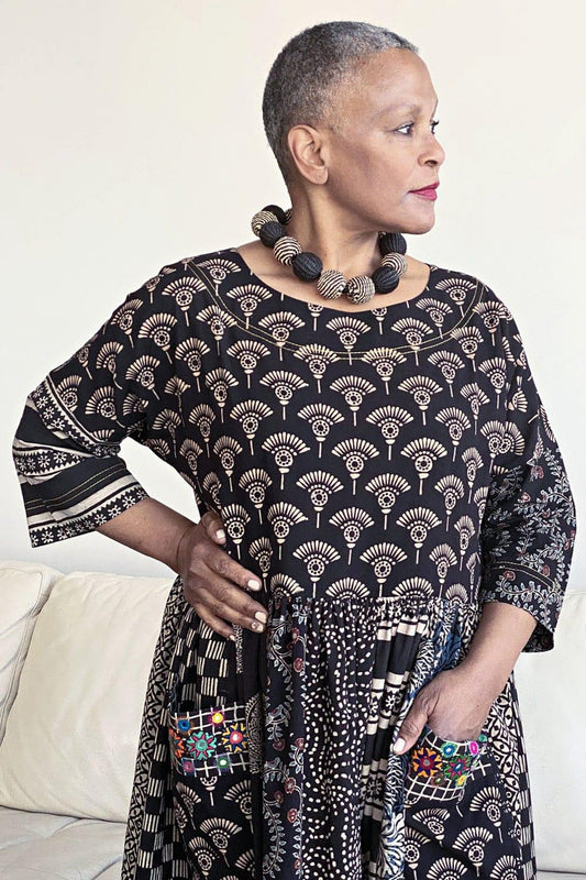 Woman with cropped grey hair looking off camera. She is wearing a woven large beaded necklace in taupe and black. She is also wearing a cotton dress handprinted with black and taupe colors.