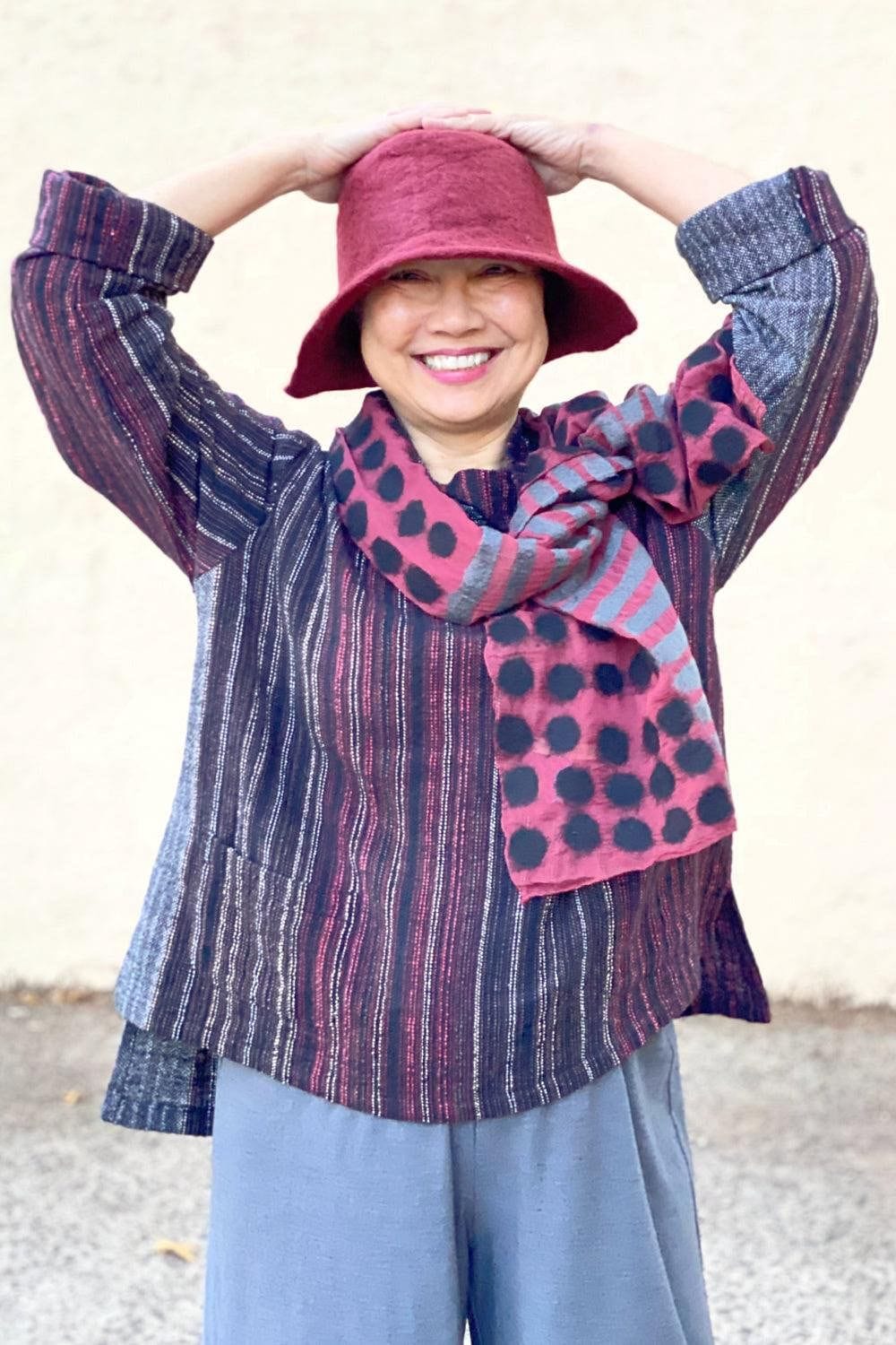 Cranberry Felted hat being worn with a matching felted scarf. A smiling woman is also wearing a handwoven pullover and grey full cut pants.