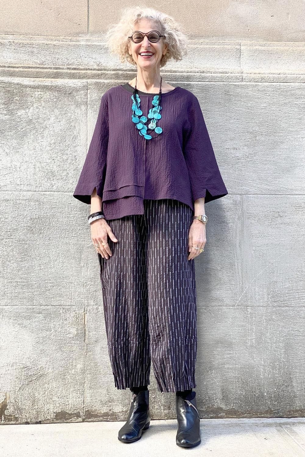 Woman over 60 wearing Plum colored  women's top with an elastic waisted loose fit black with white fleck  pant and lightwieght  statement necklace
