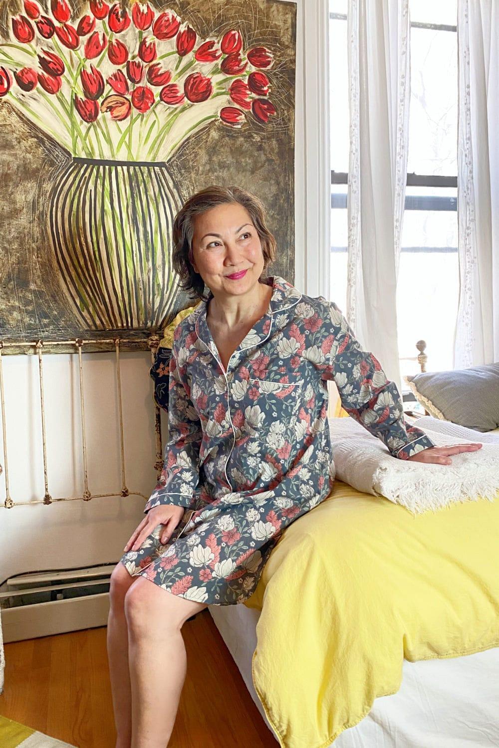 Woman sitting on the edge of a bed wearing a floral pink, grey and white night shirt. There is a floral painting behind her on the wall.
