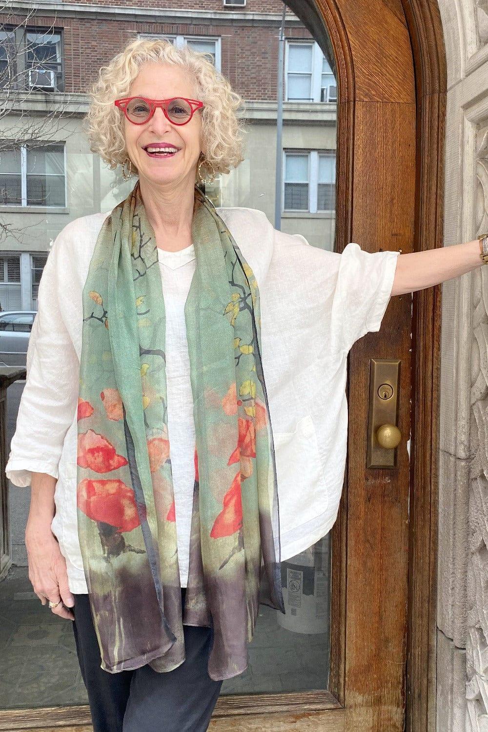 Red Blossom Print Silk Scarf draped around the shoulders of a mature woman smiling and standing infront of a doorway. She is wearing a white linen top and black cotton pants.