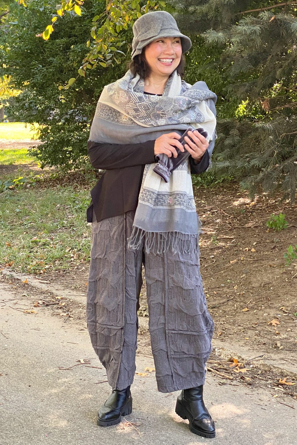 Woman standing in park wearing a fall-winter stylish outfit. A grey wool shawl, black long sleeve tee and grey loose fit textured pants with a grey bucket hat.