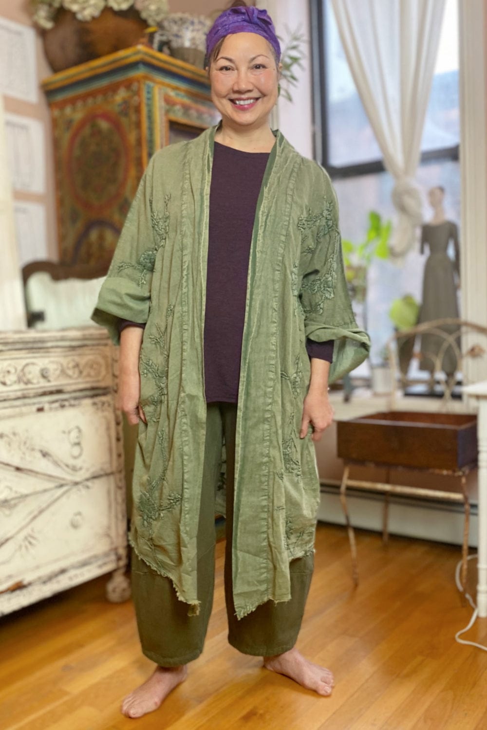 Long cotton duster jacket worn with silk scarf and linen crop pant. The outfit is green and purple and styled on a smiling woman.