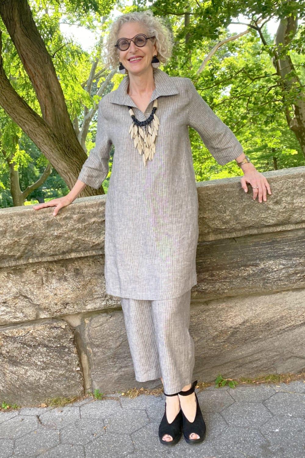 Happy woman with cropped curly hair wearing a linen long grey pinstripe tunic with matching pants. She is wearing a funky statement necklae and black open toe shoes.