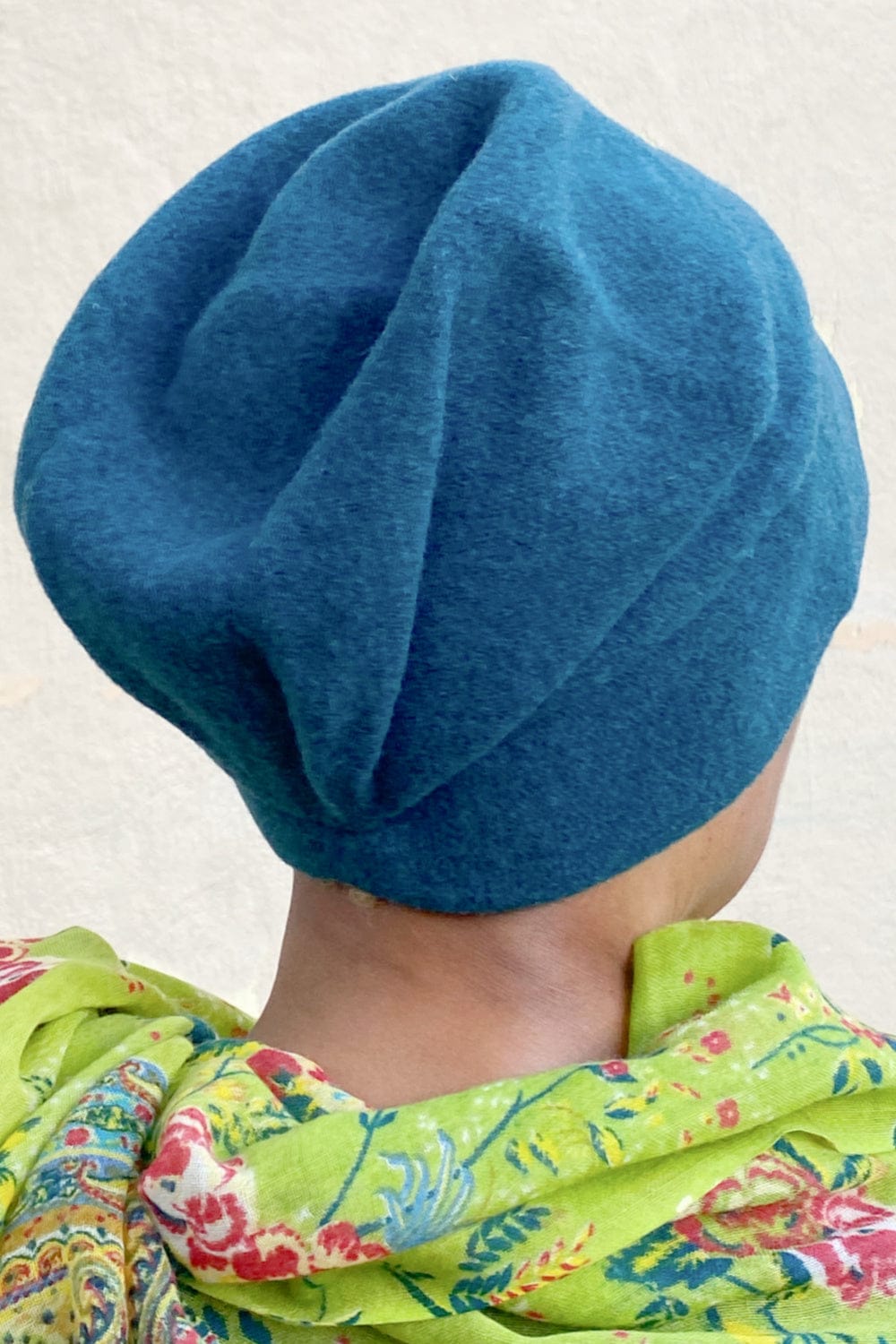 Back view of woman wearing a teal colored  merino wool  pleat hat with green floral scarf.