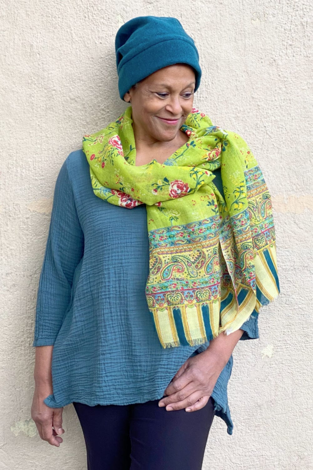 Chartreuse wool scarf with floral, paisly and stripe design worn with a dark teal winter hat on a smiling woman.