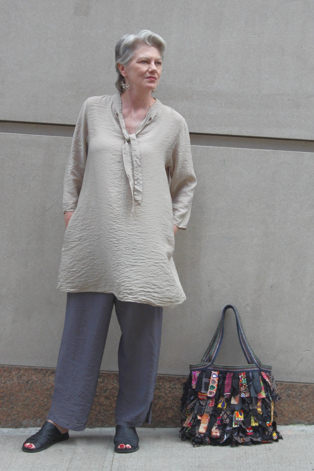 Loose cut natural color tunic with front tie worn with grey pants. The woman is standing on the street with a tote bag resting beside her.