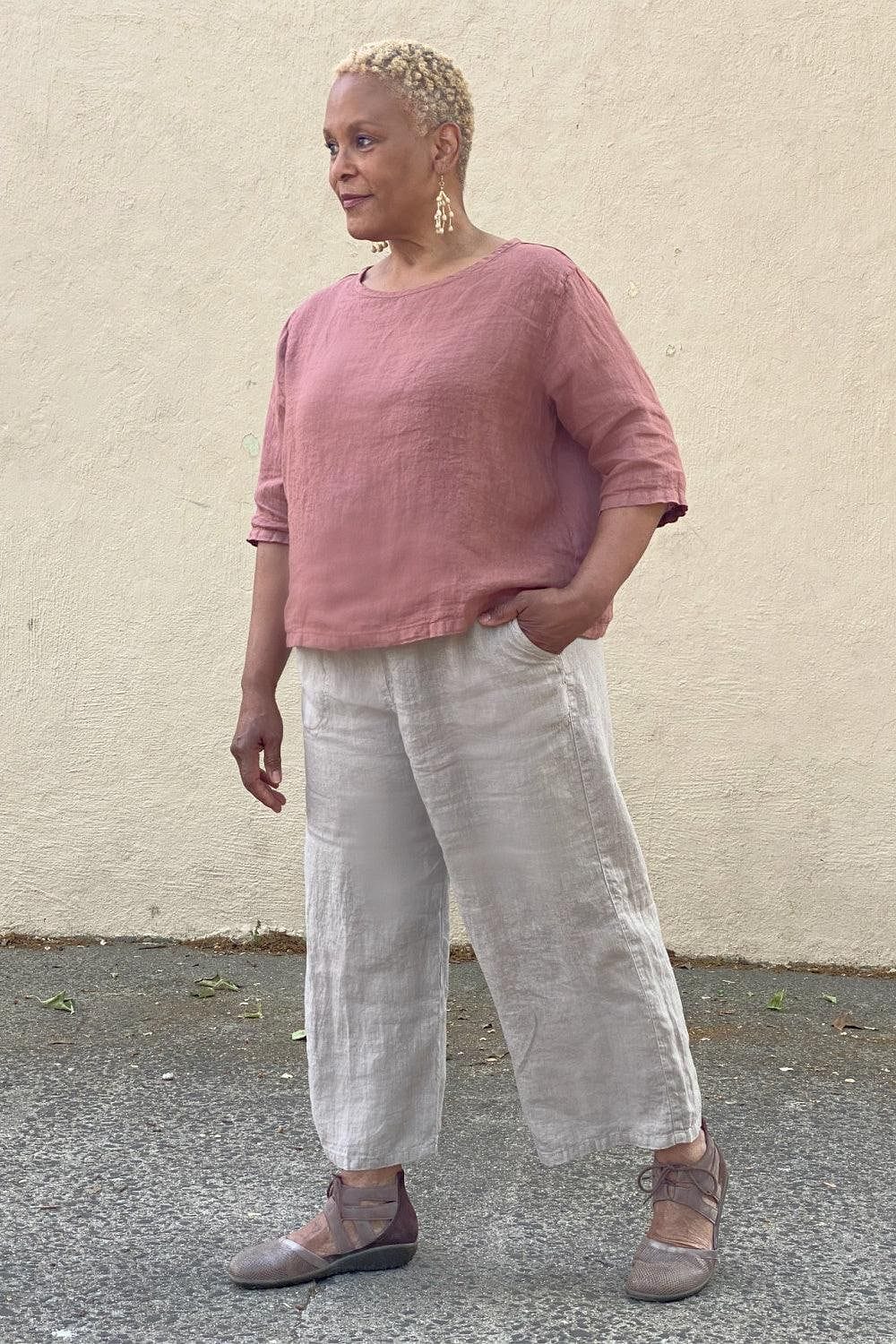 Attractive older woman with short cropped hair wearing an Elbow Sleeve Linen Top in a pretty pinkish color. She is also wearing natural color linen full cut crop pants.She is standing outside.
