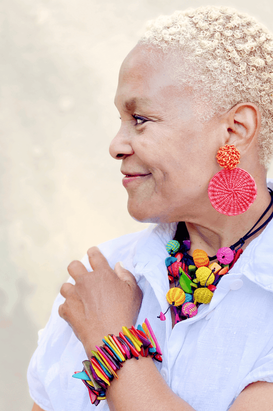 Grinning older woman with very short hair looking off in the distance. She is wearing  woven disc shaped red earrings,  bright mulit colored woven beaded necklace and taqua chipped multi colored bracelette. She is also wearing a white linen button down shirt.