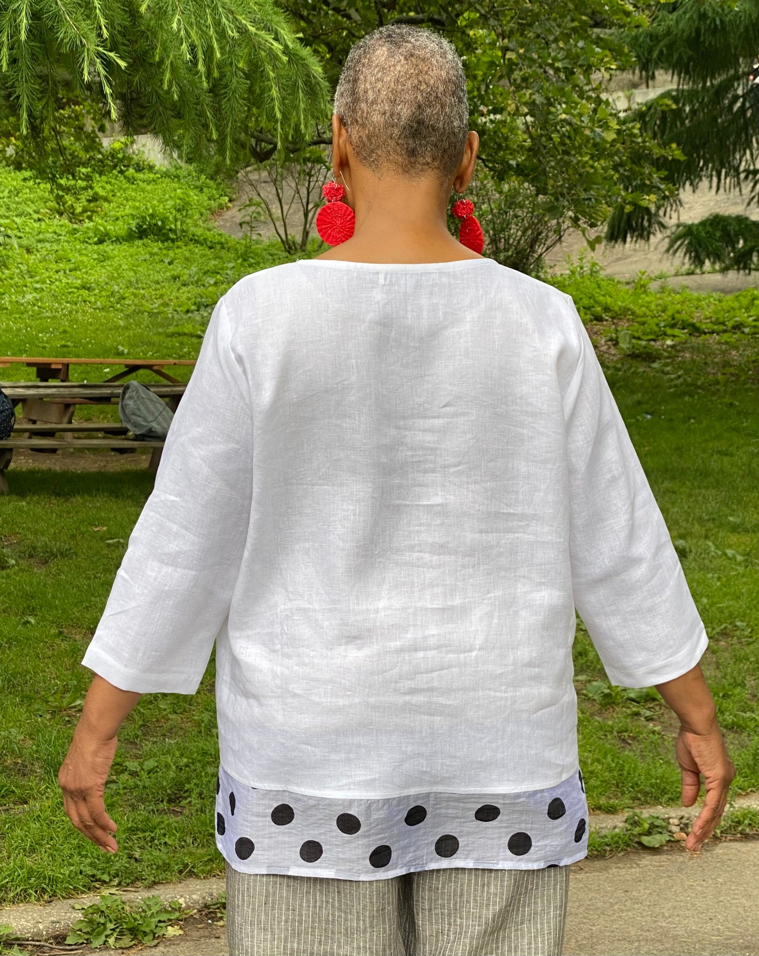 Woman with short cropped grey hair wearing a white linen top with a polka dot hem. She is showign the back view. She is wearing red woven earrings.