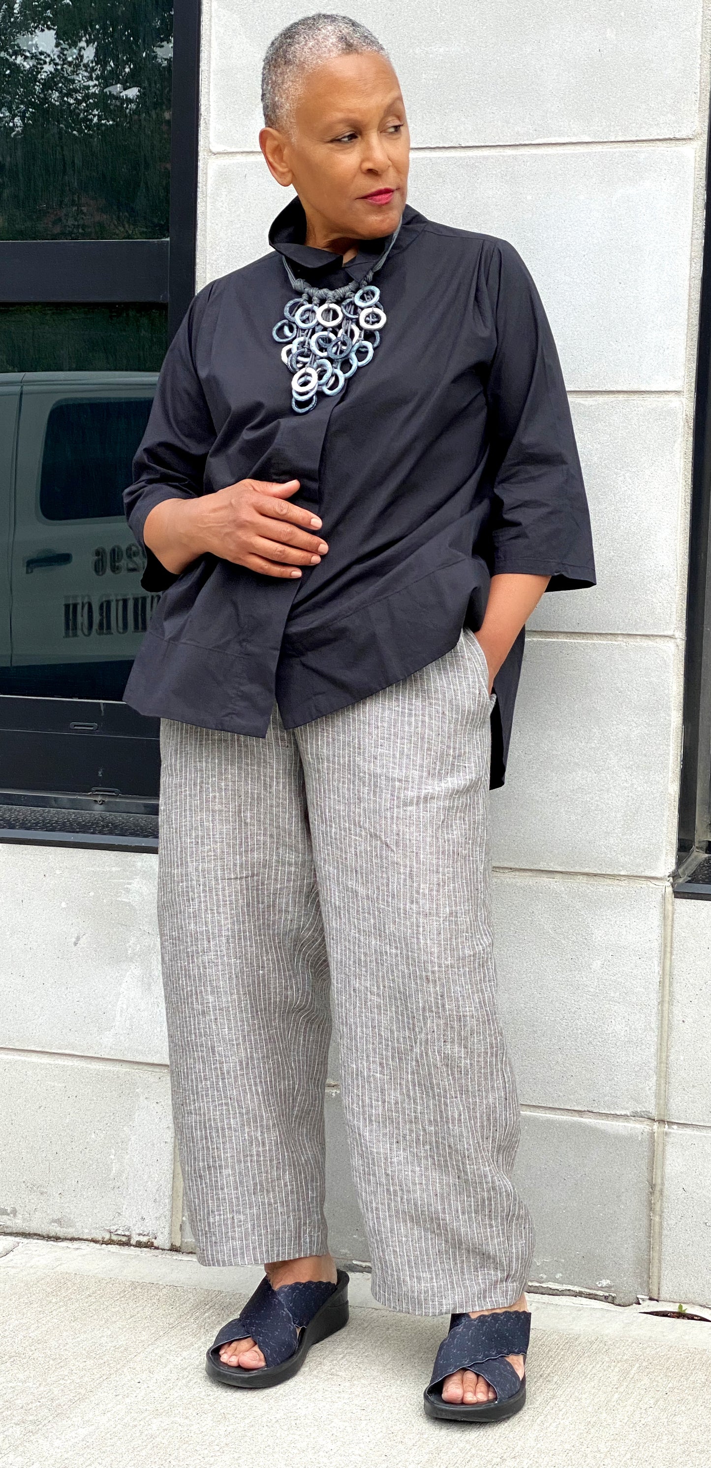 Stylish older woman stand on an city street wearing a black button down shirt, a statement necklace and a linen grey pinstripe full cut pant.