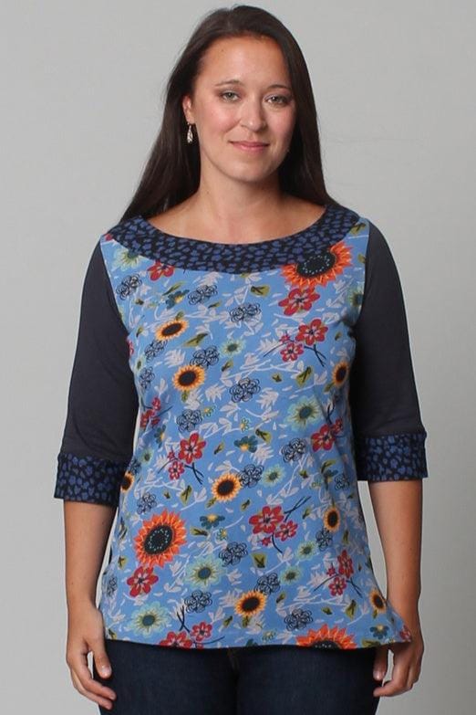 Woman standing wearing a floral print boatneck top. The front of the top has a floral print and the neck and sleeves are solid blue.