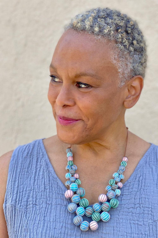 Close up of an older woman with cropped hair. She is wearing a pastel colord wooden beaded necklaces that has three layers.