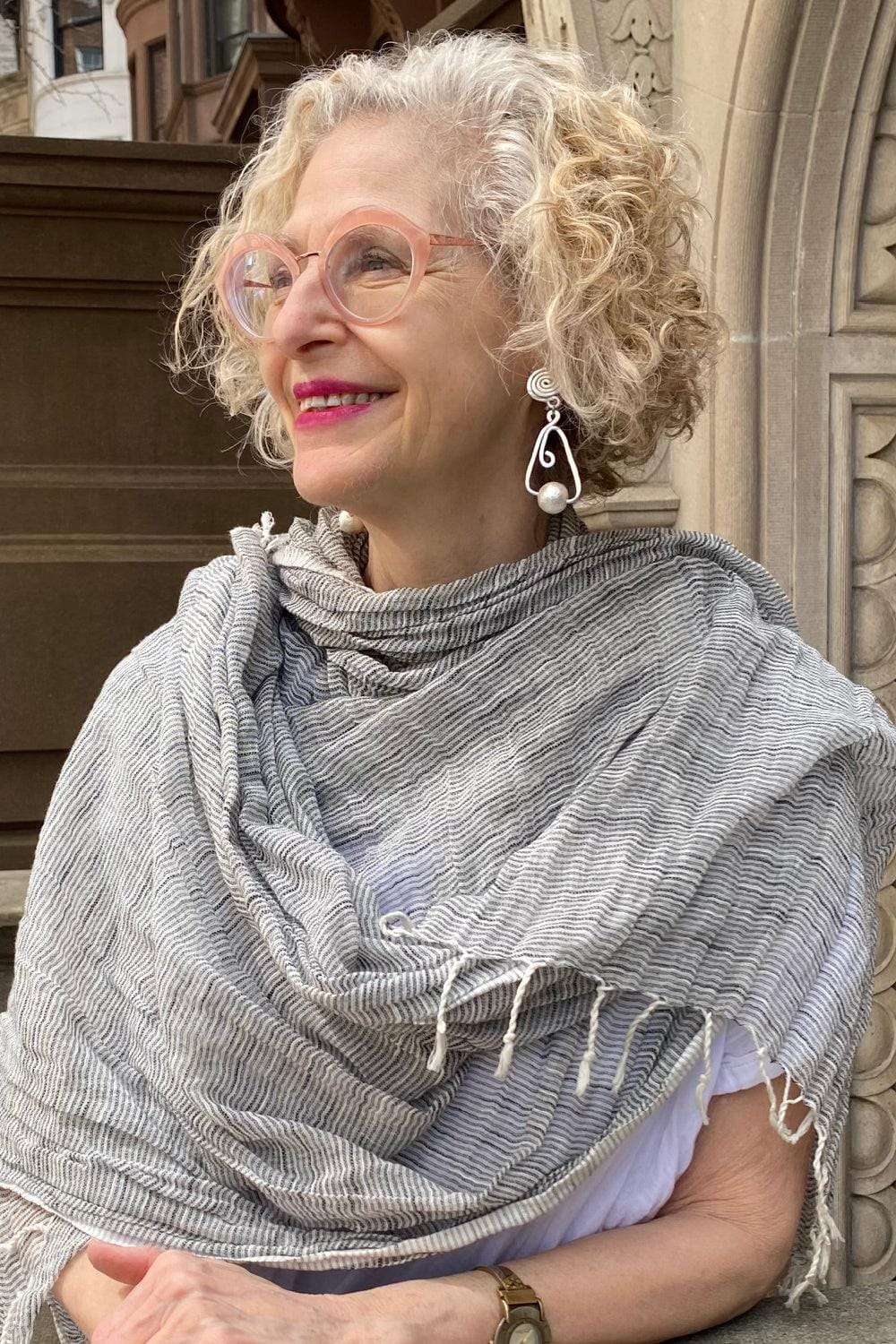 Pretty pale grey soft looking cotton scarf bing worn around the neck of an older woman with blond hair.