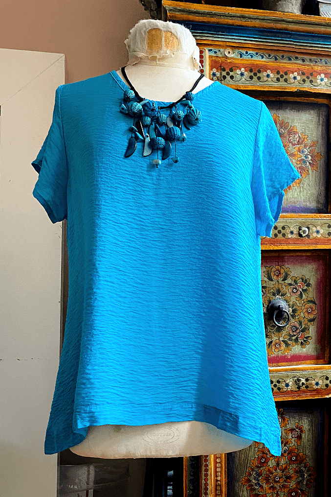 Taqua chips and handwoven beaded adjustable necklace in blue tones worn with a short sleeve blue women's tee all dresses on an old cloth mannequin standing infront of a hand painted wooden cabnet.
