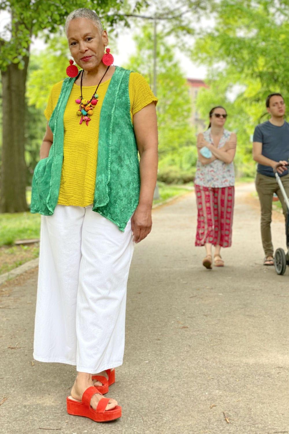 Woman wearing a mulit colored necklace with red disc earrings. She is wearing a yellow whort sleeve cotton tee with a jade green vest, white wide leg pants and red sandles.