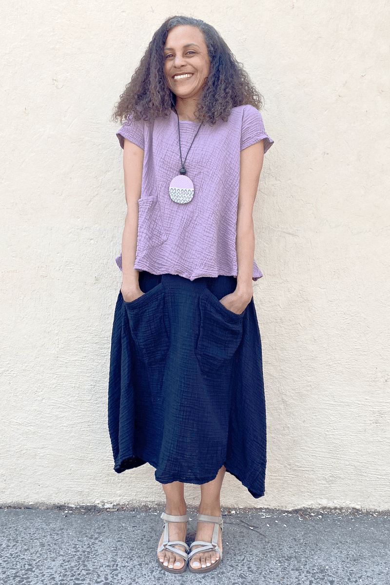 Smiling woman wearing a  Long Full cut Black Cotton Skirt with Two Front Pocket and Lots of Room. She is also wearing a lavender cotton aline cut tee and a wooden necklace in pink and black colors.