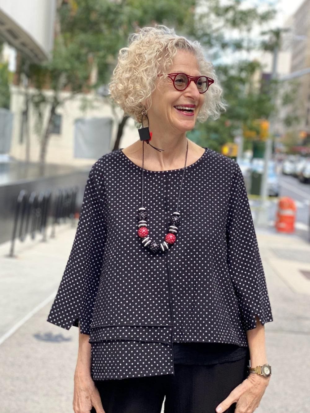 Smiling woman on the street wearing a black and white polka dot top with black and red earrings and necklace. She has blond hair and is a woman of a certain age.