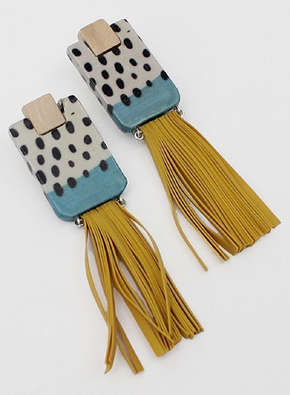 Rectangle wooden earrings with mustard colored tassels hanging from the bottom edge. The earring is colored white and blue with black dots.