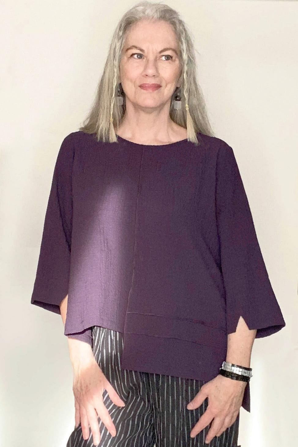 Woman with grey hair wearing a Plum colored top and black pants with a white line detail. She is looking off. She is also wearing black and clear earrings and a black and silver colored wooden wrap bracelette