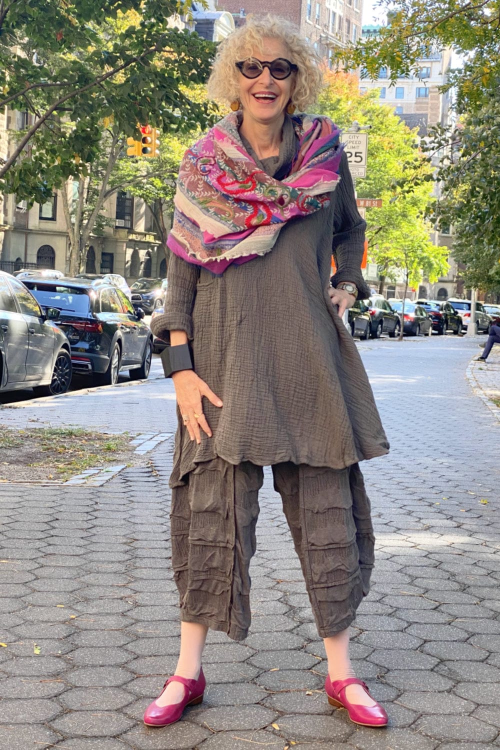 Older woman modeling on a street wearing a fushia and grey paper wool scarf styled will a grey long tunic and funky pants.