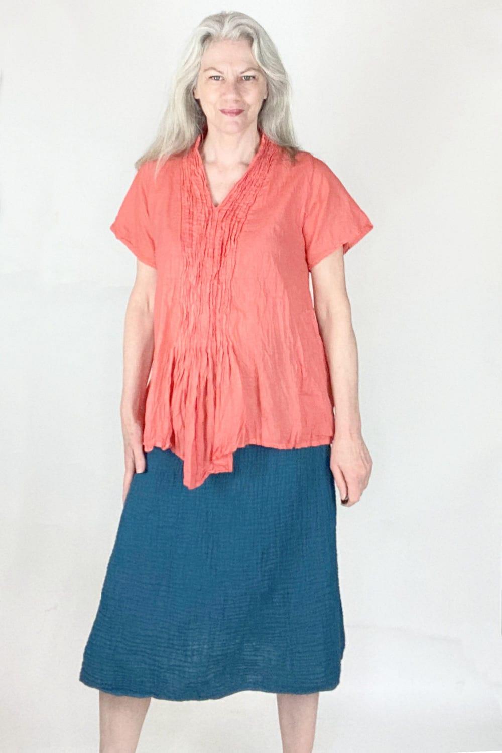 Woman with long grey hair wearing a coral coloured cotton top with short sleeves and front detail with a simple aline cut cotton navy blue skirt