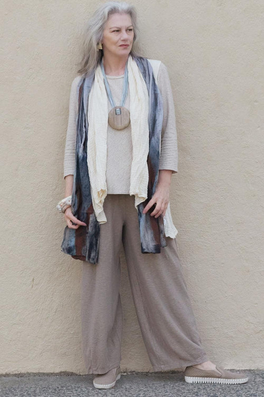 Older woman with long grey hair wearing a stylish casural outfit. She is wearing a natural colored cotton vest over a light beige tee and loose cut beige pants. Styled with a brown tone scarf and a disc pendant.