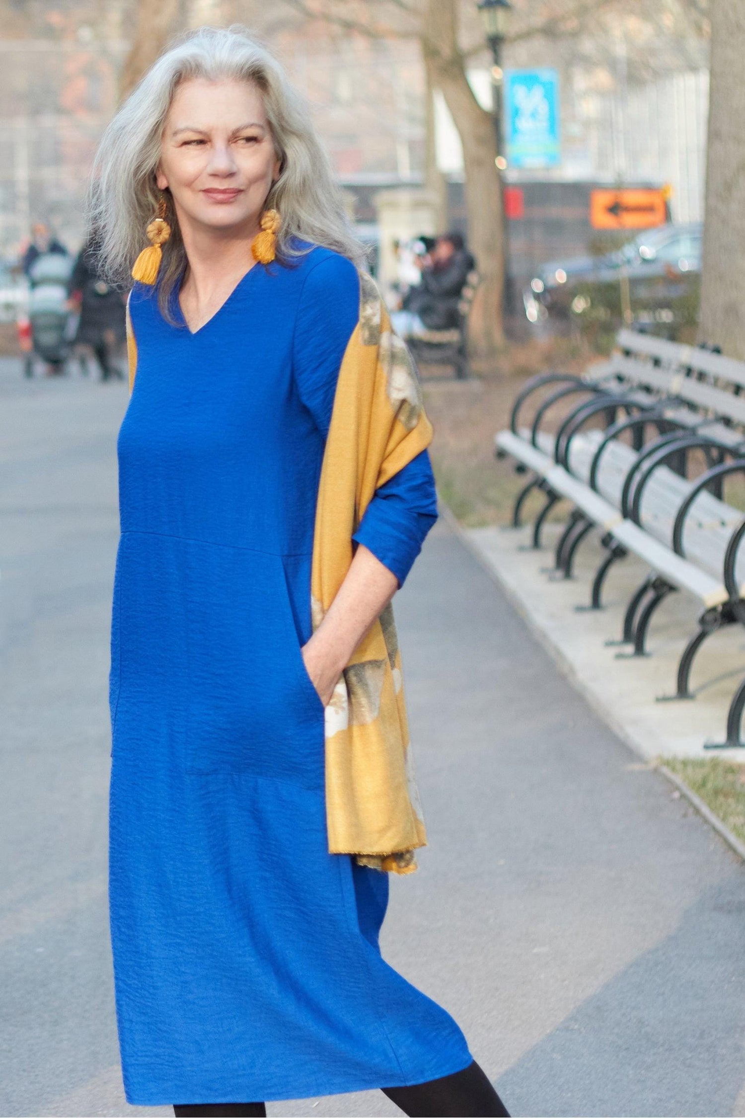 Woman with longer grey hair wearing a bright blue dress with v neckline, and yellow earrings and scarf.