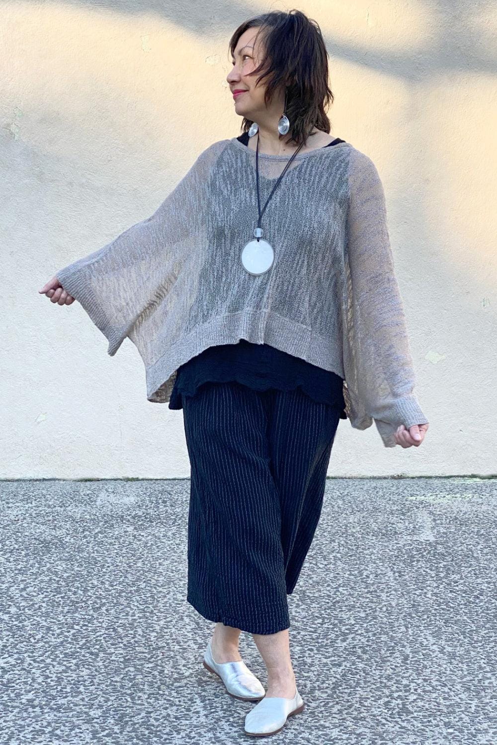 Smiling middle aged woman wearing a biege loose fit sweater with wide leg cropped pants and a pendant necklace.