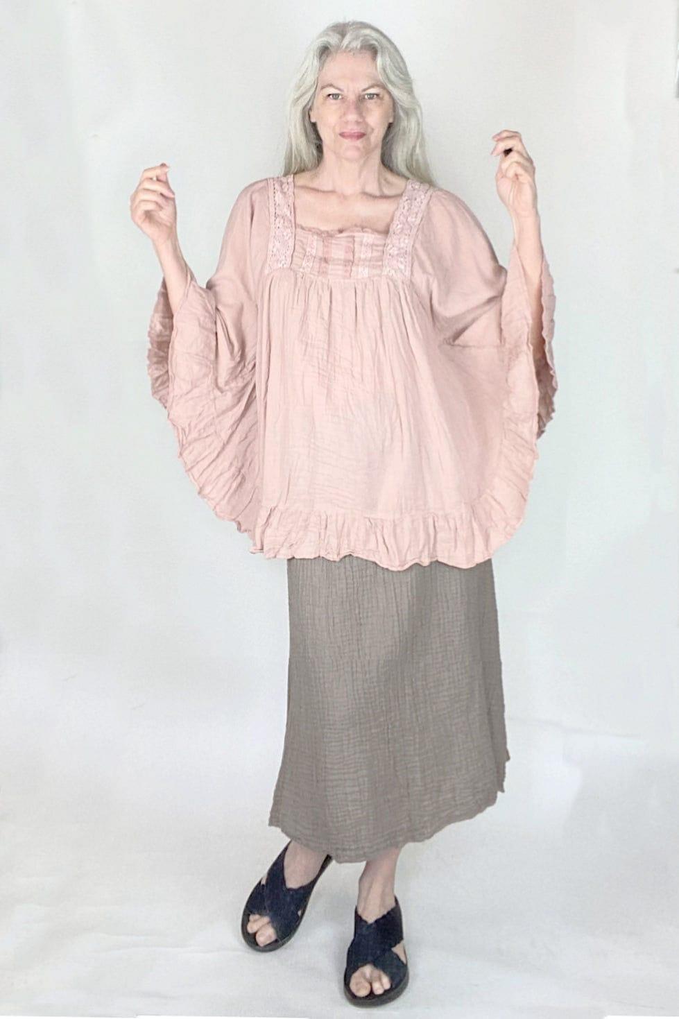 Silver haired woman modeling a pale pink cotton blouse with detail and curved ruffle hemline. Also wearing a soft beige simple cut cotton skirt.