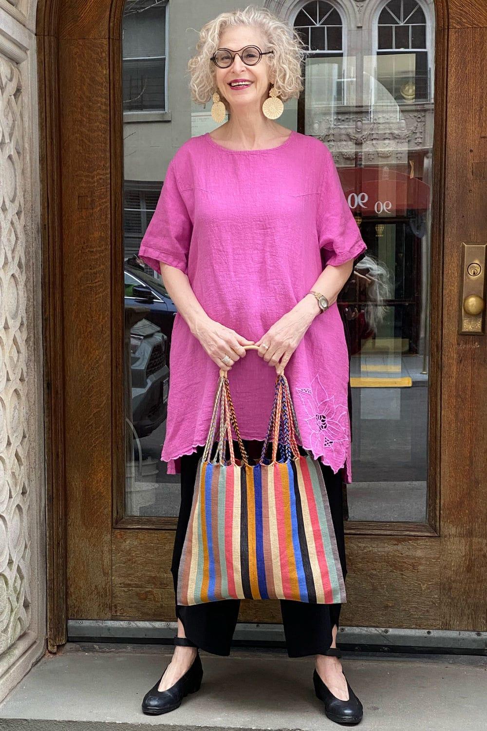 Pretty mature woman smiling and wearing loose fit casual clothing. She is holding a multi color stripe tote bag, and wearing a bright pink short sleeve linen tunic.