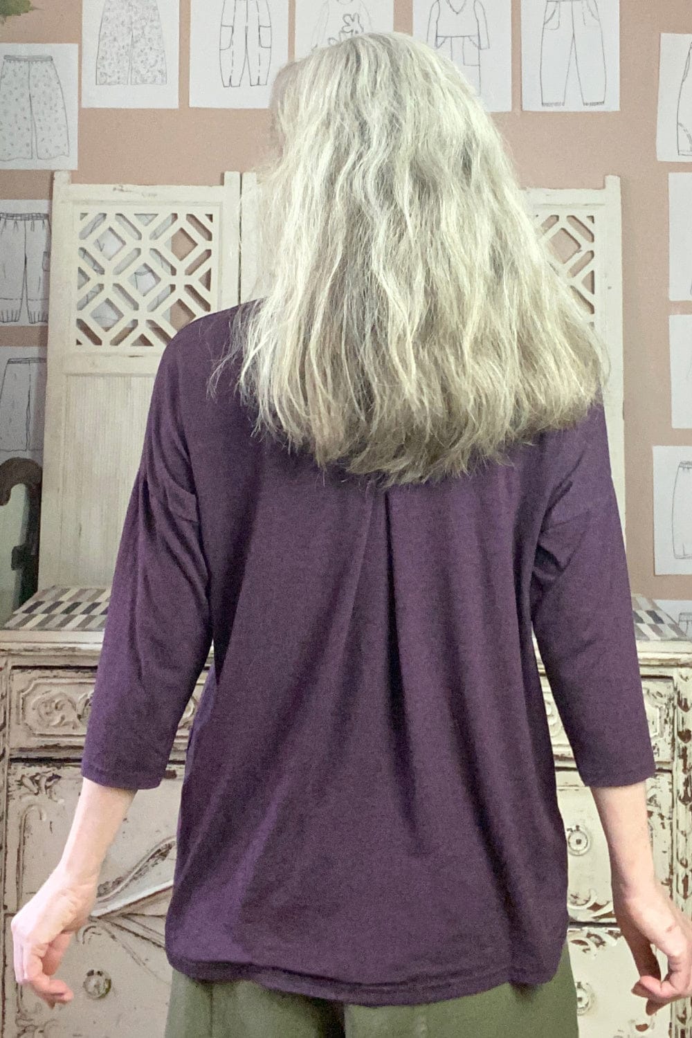 Back view of deep purple 3/4 sleeve pleated back women's tee worn on model will long grey hair.