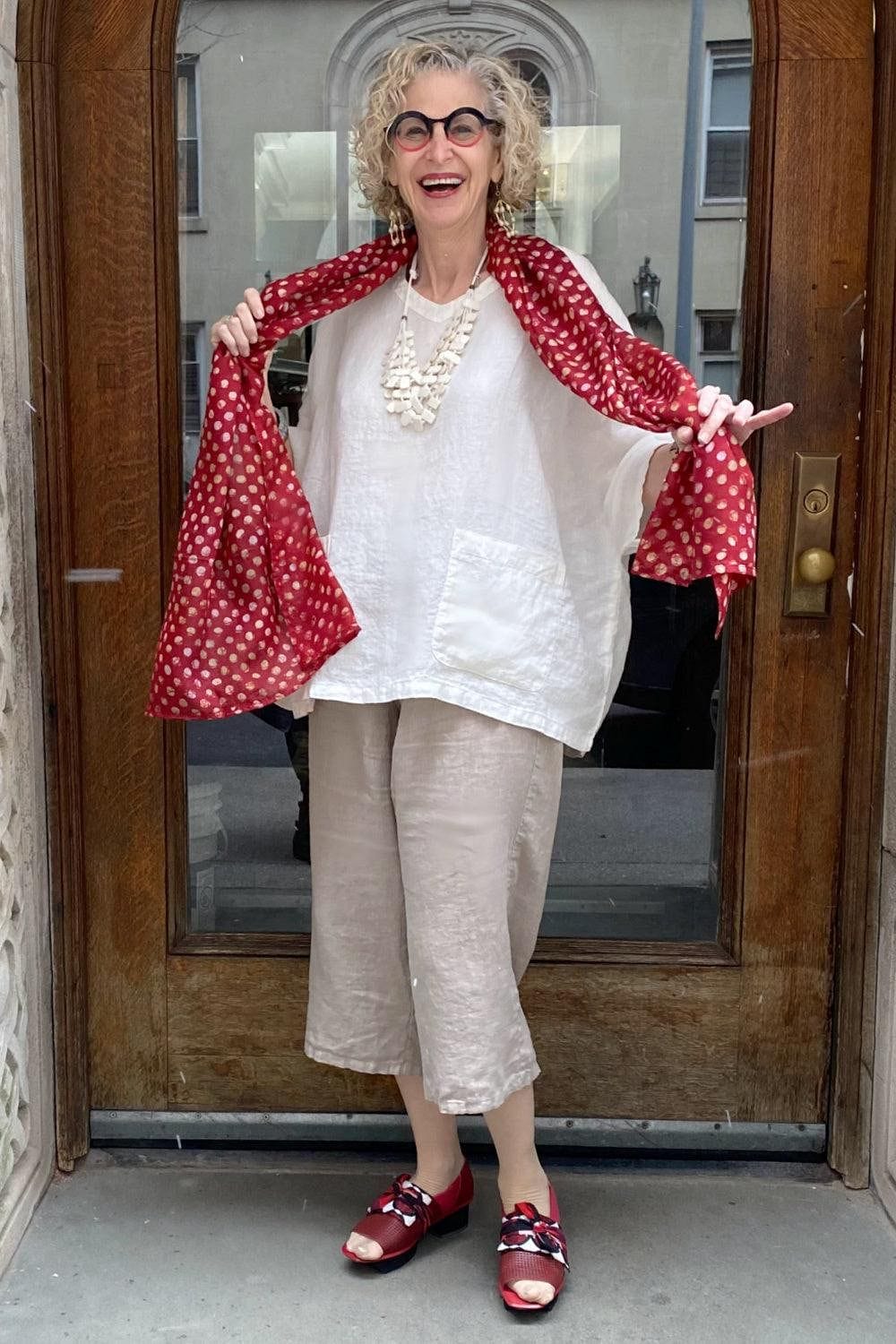 Polka Dot Silk Scarf Maroon with white dots worn over a boxy white linen tee and beige full cut linen pants. Older woman on street standing and smiling.
