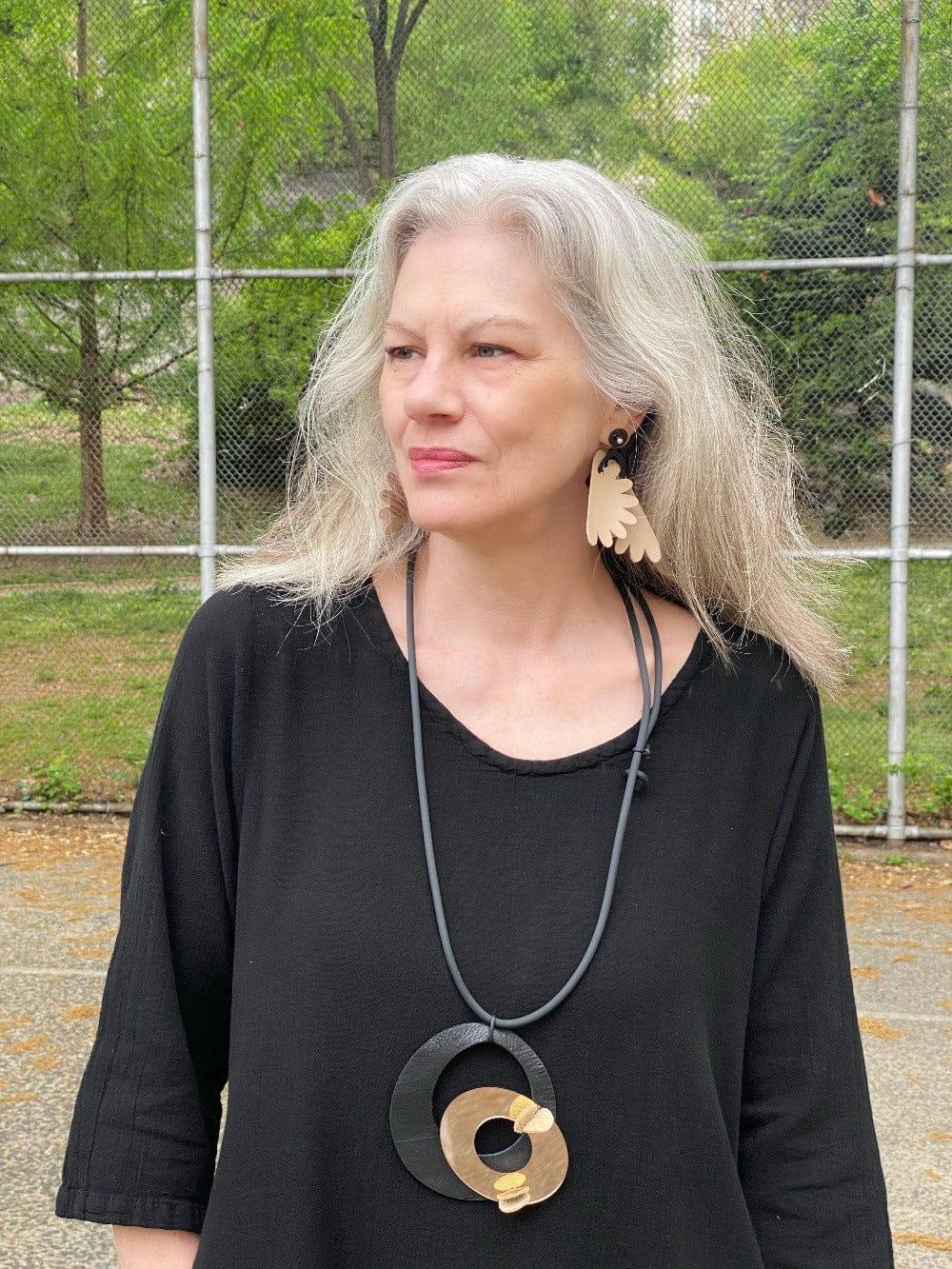 Woman with long grey hair wearing a pendant necklace with black and taupe colors. Also floral shaped taupe and black earrings.