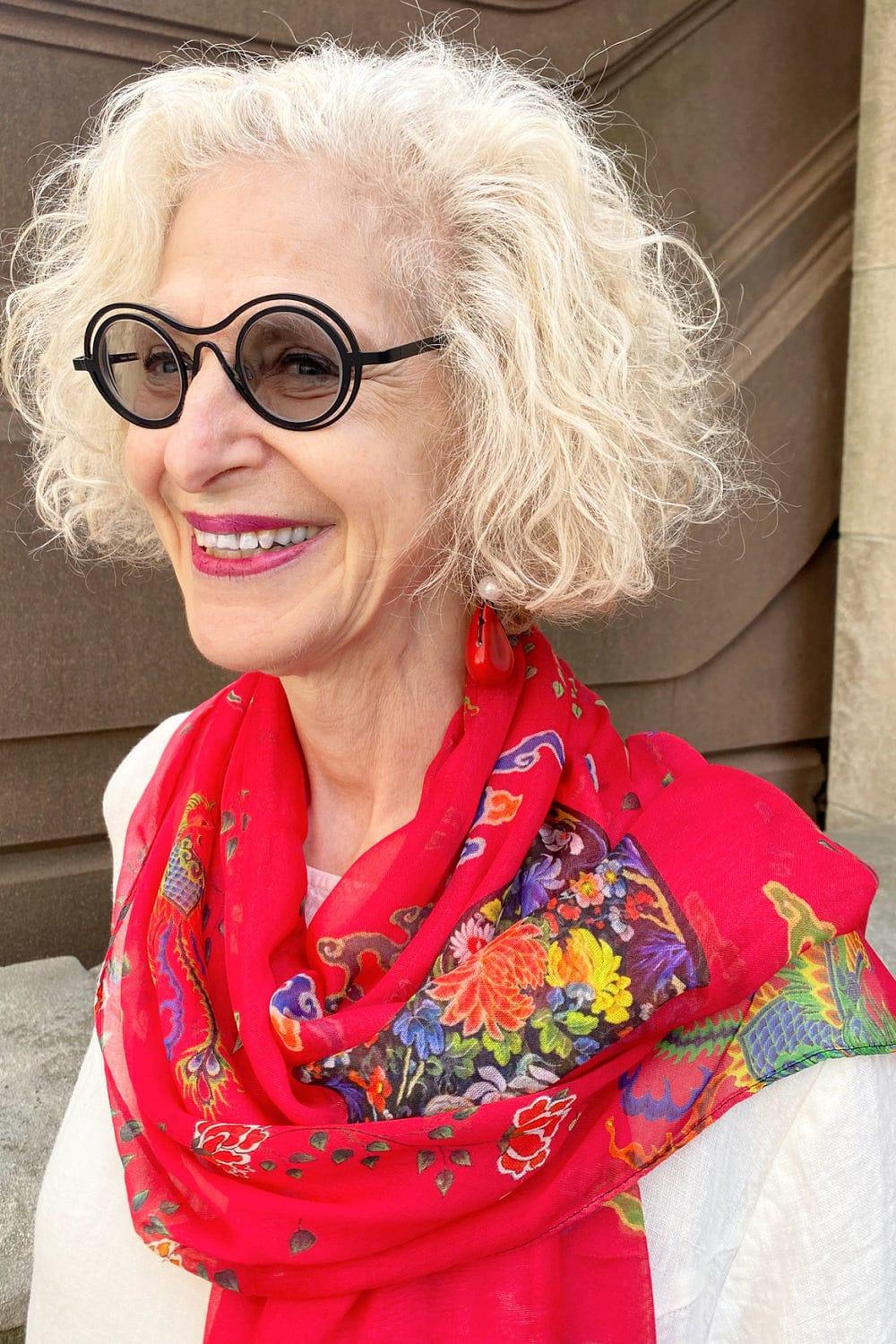 Very happy mature woman wearing a beautiful red drapy scarf over her shoulders. She is wearing red drop earrings and a white linen top with black rim glasses.